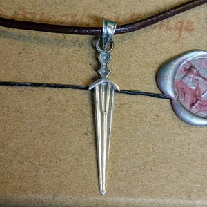 May include: A silver pendant in the shape of a dagger hangs from a brown leather cord. The pendant has a detailed design with a pointed tip and a slightly curved blade.