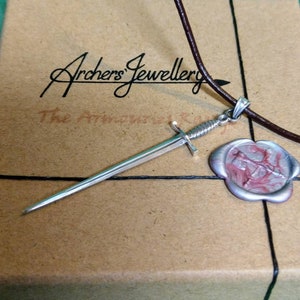 May include: A silver sword pendant necklace with a brown leather cord. The pendant is on a brown box with the text "Archers' Jewellery" and "The Armoured King".