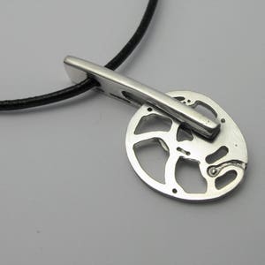 Può includere: Una collana con un ciondolo in argento su un cordoncino in pelle nera. Il ciondolo è di forma circolare con un intricato motivo traforato.