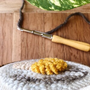 May include: Close-up of a round rug with a circular pattern of gray, white, and beige yarn. A small section of the rug is a bright yellow color. A wooden needle with a metal hook is lying on a wooden surface.