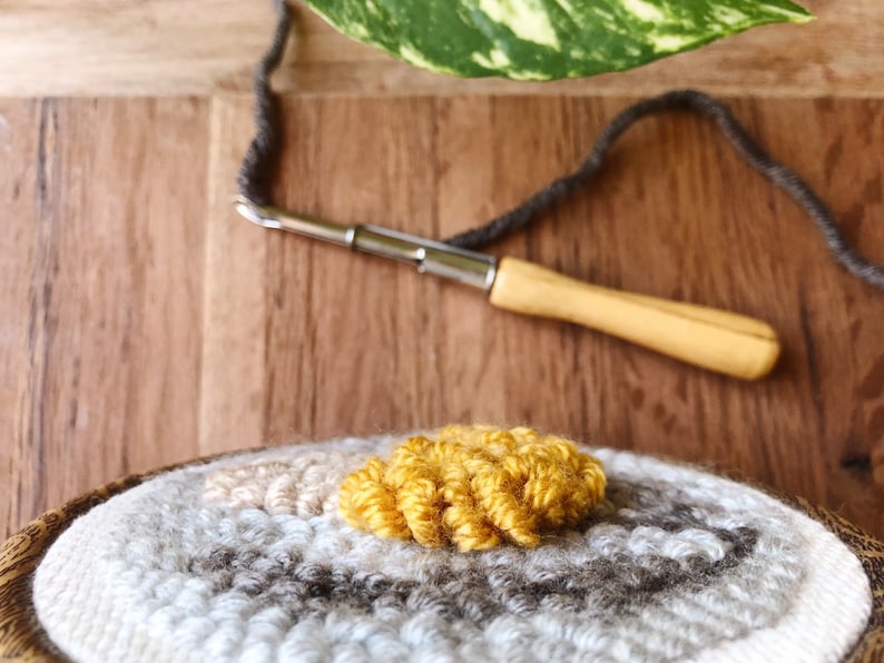 May include: Close-up of a round rug with a circular pattern of gray, white, and beige yarn. A small section of the rug is a bright yellow color. A wooden needle with a metal hook is lying on a wooden surface.
