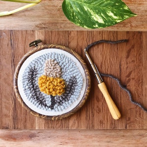May include: A close-up of a round, gray, brown, and yellow wool rug with a wooden frame. The rug is being worked on with a wooden needle and gray yarn.