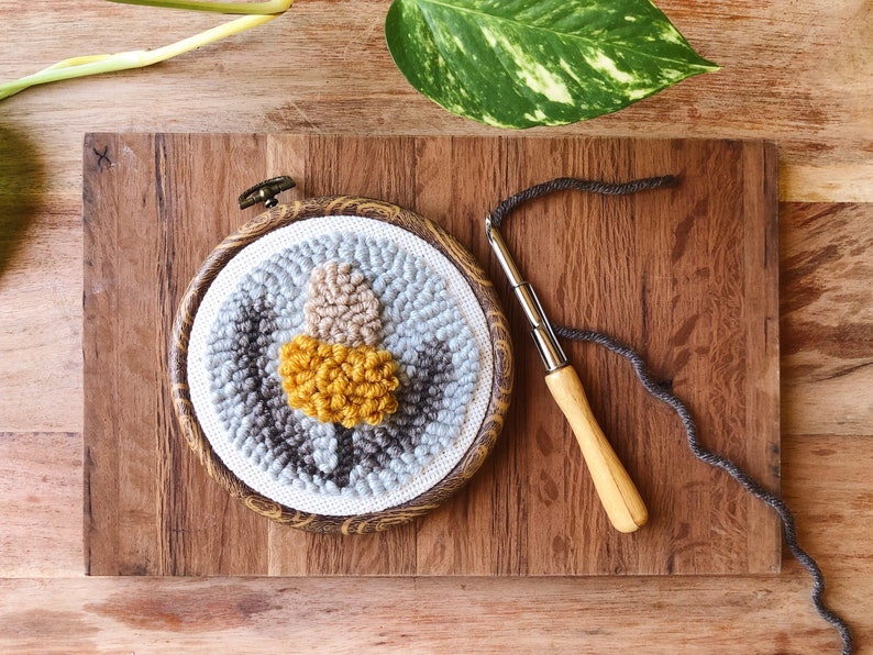 May include: A close-up of a round, gray, brown, and yellow wool rug with a wooden frame. The rug is being worked on with a wooden needle and gray yarn.