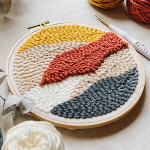 Op de afbeelding: Een ronde borduurring met een kleurrijk abstract ontwerp gemaakt met garen en een punchnaald. Het ontwerp heeft tinten geel, beige, oranje en grijs.
