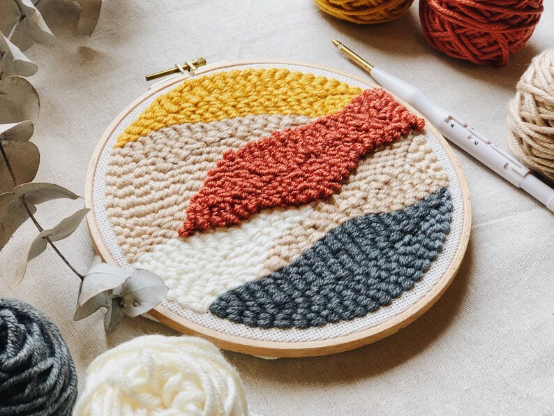 Op de afbeelding: Een ronde borduurring met een kleurrijk abstract ontwerp gemaakt met garen en een punchnaald. Het ontwerp heeft tinten geel, beige, oranje en grijs.