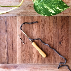 May include: A wooden crochet hook with a brown yarn on a wooden surface. The hook has a wooden handle and a metal hook. The yarn is coiled on the surface.