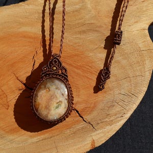 May include: A handmade macramé necklace with an oval pendant made of cream and tan stone. The pendant is intricately woven with brown cord, and the necklace rests on a wooden surface. The necklace has a braided cord.