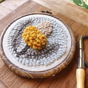 May include: A close-up of a round, wooden-framed embroidery hoop with a colorful wool punch needle design. The design features three circular shapes in shades of gray, beige, and yellow.