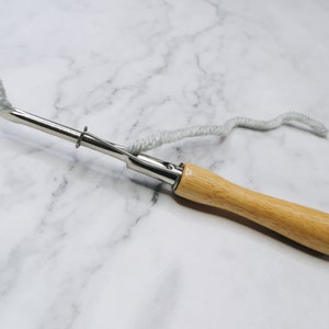 May include: A wooden handled yarn needle with a metal hook and a loop at the end of the handle. A length of white yarn is threaded through the needle.