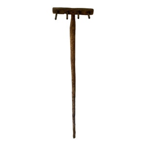 Pode incluir: Um ancinho de madeira com um cabo longo e cinco dentes de metal. O ancinho é feito de madeira natural e tem um aspecto rústico.