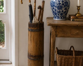 Puede incluir: Un paragüero cilíndrico de madera alto, con tablones verticales desgastados y bandas de metal oscuro. Contiene varios paraguas con mangos variados, incluyendo un paraguas negro y uno de color claro, colocados en posición vertical.