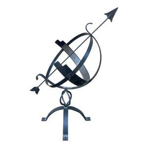 May include: A black metal sundial with an arrow-shaped gnomon. The sundial features a spherical design with Roman numerals and is mounted on a decorative base with three curved legs. The arrow points towards the sky.