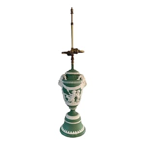 May include: A green ceramic table lamp with a white relief design of figures and a Greek key pattern. The lamp has a brass base and a white shade.