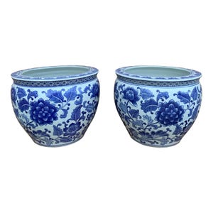 May include: Two matching blue and white ceramic planters. Each planter features a floral design with blue flowers and leaves on a white background. The planters have a round shape with a wide opening.