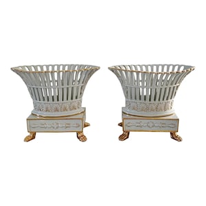 Può includere: Coppia di cesti in ceramica bianca con lati traforati e finiture dorate. Ogni cesto poggia su una base quadrata con dettagli dorati e piedi a zampa. Design classico ed elegante.