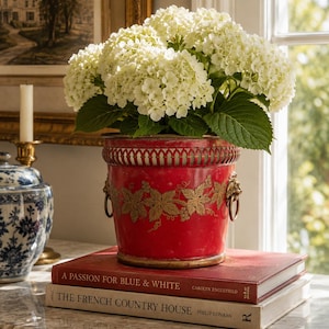 May include: A vibrant red metal planter with a gold-toned grape vine motif and lion head ring handles, containing a large bouquet of white hydrangea flowers. The planter rests on two stacked hardcover books with visible text on their spines.