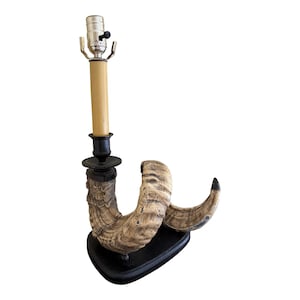 Pode incluir: Uma lâmpada de mesa única com uma base de chifre de carneiro curvada sobre uma plataforma de madeira preta. A lâmpada tem uma coluna bege e um soquete prateado. O chifre tem uma textura em tons de castanho claro e creme.