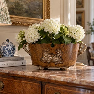 May include: An oval-shaped brown planter with a mottled finish, featuring two side handles and a central relief ornament of leaves and a bow. The planter stands on four small feet and is filled with a large cluster of white hydrangea flowers.