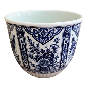 May include: A white ceramic cup featuring a deep blue floral pattern arranged in vertical panels. The design includes intricate botanical illustrations and decorative borders around the exterior, with a smooth, rounded rim and a fluted base.