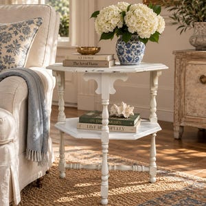 May include: A white hexagonal side table with a distressed finish, featuring three turned legs and a lower shelf. The top surface holds a blue and white floral vase with white hydrangeas, a gold bowl, and two books titled At Home and The Art of Home.