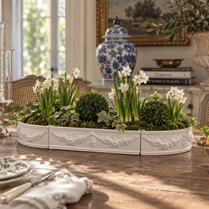 May include: A white ceramic planter with three connected sections featuring embossed floral garland details. The planter is filled with green moss, two round boxwood topiaries, and three clusters of white narcissus flowers with green stems.