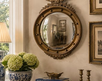 Pode incluir: Um espelho convexo redondo em tom dourado com um emblema decorativo de águia no topo e uma série de pequenas esferas ao redor da moldura interna. O espelho está montado em uma parede acima de um console de madeira.