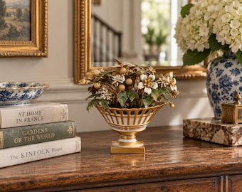 May include: A white and gold porcelain footed bowl with a lattice rim design, filled with a decorative arrangement of artificial white flowers, green leaves, and small brown seed pods, resting on a wooden surface next to a stack of three books.