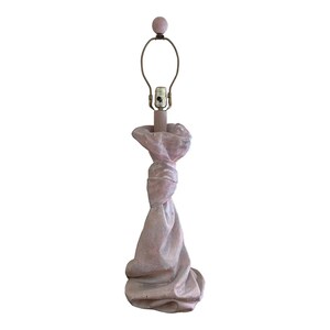 May include: A pink ceramic table lamp shaped like a knotted bag. The lamp has a simple metal shade and a round finial.