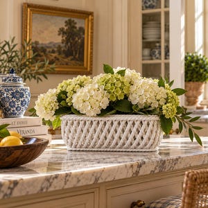 May include: A rectangular white ceramic basket with a woven rope-like texture filled with a large arrangement of white and light green hydrangea flowers and green leaves, positioned on a marble surface.