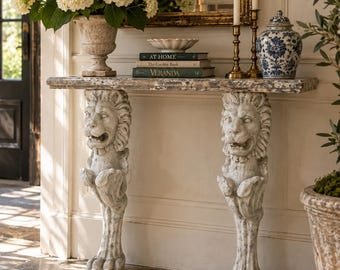 May include: A console table featuring a rectangular stone top supported by two carved stone lion pedestals. The table is decorated with a stone urn filled with white hydrangeas, a stack of three books titled At Home, The Garden Book, and Veranda, two brass candlesticks, and a blue and white porcelain jar.