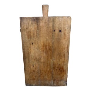 May include: A rectangular wooden cutting board with a light brown, weathered grain and a prominent vertical handle at the top center. The surface displays visible wood texture, natural knots, and signs of wear across the flat, sturdy board.
