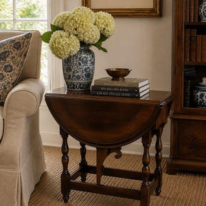 May include: A dark wood drop-leaf table with turned legs and a central support. On the table sits a blue and white floral vase filled with white hydrangea flowers, a small brass bowl, and three stacked books titled Houses, The English Room, and A Place to Call Home.
