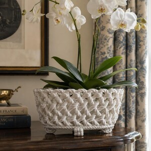 May include: A white ceramic planter with a woven rope-like texture and a rectangular shape, featuring four small feet at the base. A white orchid plant with green leaves is potted inside the planter, which sits on a dark wooden surface.