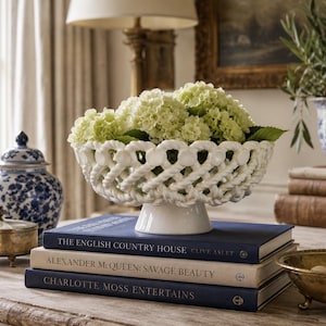 Peut inclure: Un bol sur pied en céramique blanche au design en treillis tressé, rempli de fleurs d'hortensia vertes. Le bol repose sur une pile de trois livres intitulés The English Country House, Alexander McQueen: Savage Beauty et Charlotte Moss Entertains.