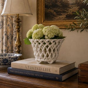 May include: A white ceramic basket-weave bowl filled with light green hydrangea flowers, resting on a stack of two hardcover books titled A Visited Life and The English Country House. The bowl features a lattice design and small feet.