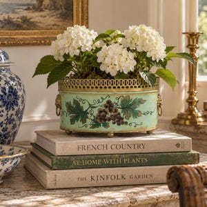 Pode incluir: Um vaso oval de metal verde-claro com uma borda dourada recortada e alças em forma de cabeça de leão. O vaso apresenta um design pintado de uvas castanhas e folhas verdes, preenchido com flores de hortênsia brancas e folhagem verde.