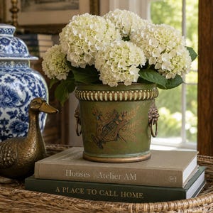 May include: A green metal planter with a gold-toned rim and lion head ring handles, featuring a black graphic of a lyre and foliage. The planter is filled with a large bouquet of white hydrangea flowers and sits atop two stacked hardcover books.