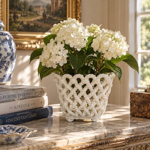 May include: A white ceramic planter featuring a woven rope-like lattice design, containing a cluster of white hydrangea flowers with green leaves. The planter has a flared rim and a small footed base, providing a textured look for floral displays.
