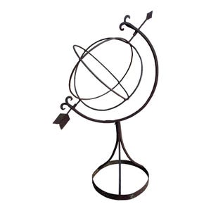 May include: A decorative metal armillary sphere with a dark brown, weathered finish. The sphere features intersecting rings and is mounted on a stand with a circular base. An arrow design is incorporated into the structure.