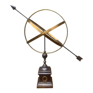May include: A bronze and brass armillary sphere. The sphere features a circular frame with intersecting bands, an arrow, and a decorative base with a laurel wreath. The arrow extends through the sphere.