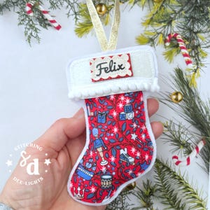 May include: A small fabric Christmas stocking ornament featuring a red background with a pattern of blue toys, including a nutcracker, drum, and blocks. The white cuff has a scalloped tag with the name Felix in black script and a gold ribbon loop.