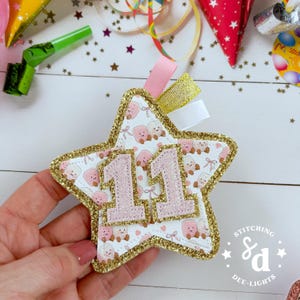May include: A star-shaped ornament with a white background featuring a pattern of small pink marshmallows and bows. The star is outlined in gold glitter and features the number 11 in large, light pink glittered digits. Pink, gold, and white ribbons.