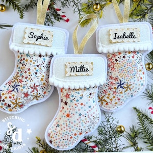 May include: Three small fabric Christmas stockings featuring a colourful star pattern on a white background. Each stocking has a white cuff with pom-pom trim, a gold glitter hanging loop, and a rectangular name tag reading Sophie, Millie, or Isabelle.