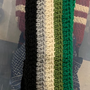 Puede incluir: Una diadema de crochet con rayas negras, grises, blancas, verdes y verde oscuro.