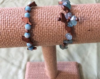 May include: Two bracelets with blue and green beads and copper bird charms displayed on a burlap jewellery stand.