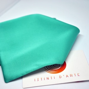 Può includere: Un tessuto piegato di colore turchese, possibilmente seta o raso, è il punto focale di questa immagine. Una carta bianca con il testo "ISTINTI D'ARTE" in lettere arancioni e rosse è parzialmente visibile, suggerendo un marchio o un'etichetta di prodotto.