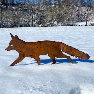May include: A rusty metal fox silhouette standing in a snowy field. The fox is facing left and has a long tail. The background is a snowy field with trees in the distance.
