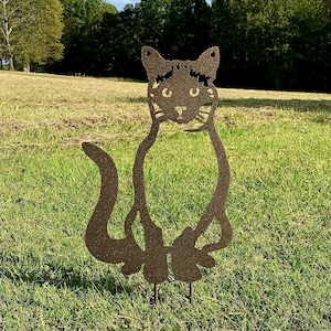 May include: A metal cat garden stake with a brown finish. The cat has a stylized design with cut-out details for the eyes, nose, and mouth. The cat is sitting upright with its tail curled. The stake is placed in a grassy field with trees in the background.