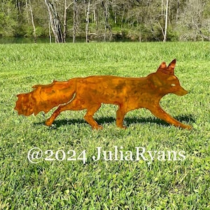 May include: A rusty metal fox sculpture standing in a grassy field. The fox is facing right and has its tail curled behind it. The sculpture is in a field with green grass and a blue sky in the background. The text "@2024 Julia Ryans" is visible in the bottom center of the image.