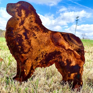 K&ouml;nnte beinhalten: Eine rostige Metallsilhouette eines Hundes, der auf einem Bett aus trockenem Gras steht. Der Hund ist nach rechts gerichtet, mit dem Kopf nach oben geneigt. Die Silhouette ist eine einfache Umrandung der Hundeform, ohne Details.